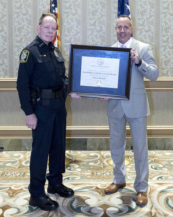 A police officer and a gentleman smile as they hold a large, framed award.