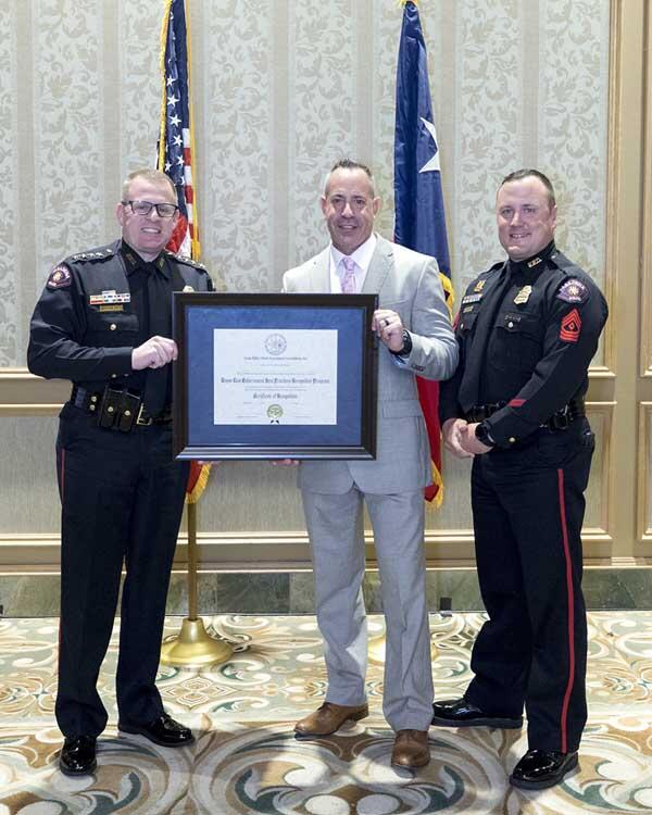Two police officers stand smiling on either side of a gentleman, holding a large, framed award. 