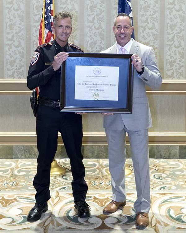 A police officer smiles brightly as he and a gentleman hold a large, framed award. 