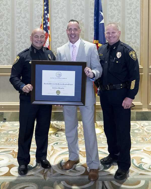 Two officers and a gentleman stand smiling as they hold a framed award. 