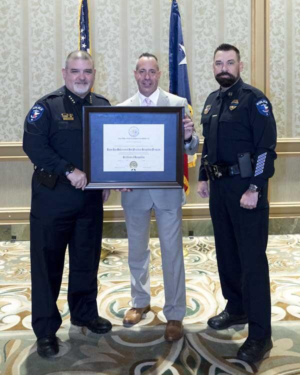 Two officers stand on either side of a gentleman who is holding a large, framed award. 