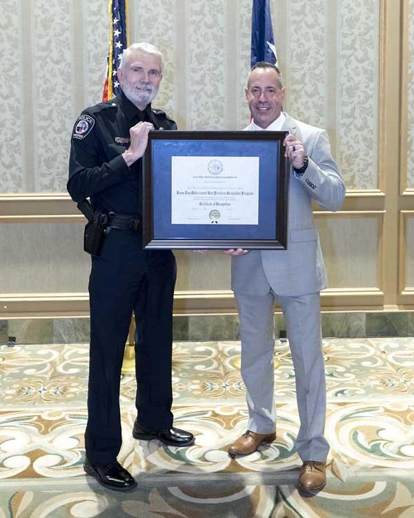 A police officer and a gentleman smile as the stand on either side of a large, framed award.