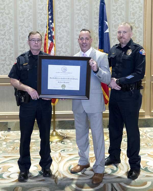 Two police officers stand on either side of a gentleman holding a large, framed award