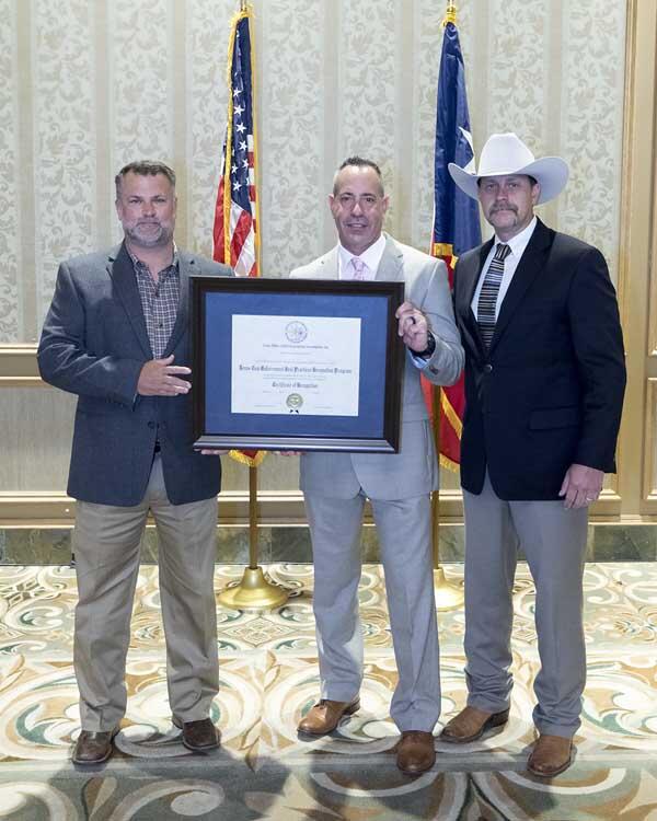 Three gentlemen, one wearing a cowboy hat, stand together holding a large, framed award.