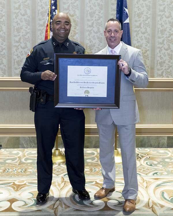 A police officer and a gentleman stand together smiling as they hold a large, framed award.