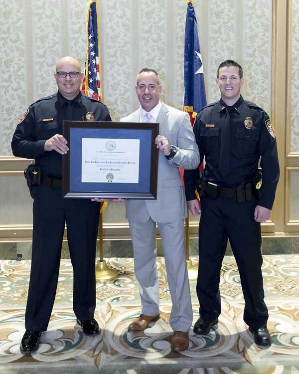 Two smiling police officers stand on either side of a gentleman, holding a large, framed award.