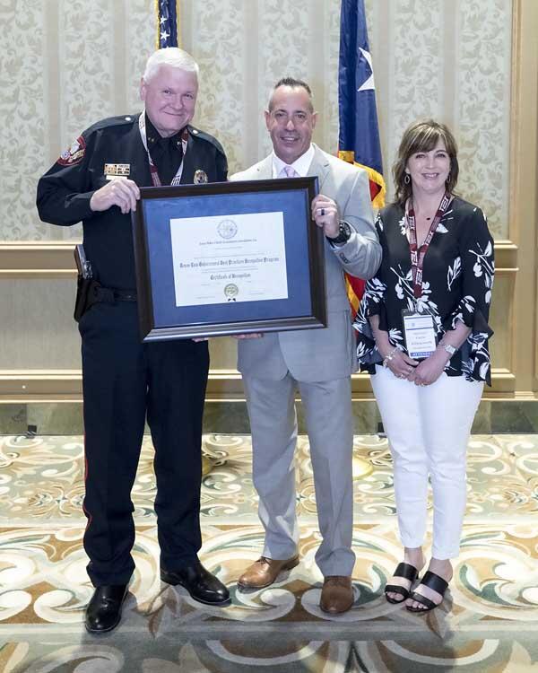 A woman stands with a gentleman and a police officer that are smiling and holding a large, framed award.