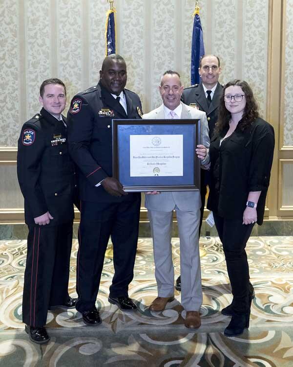 Two police officers, two gentleman, and a woman stand together holding a large, framed award. 