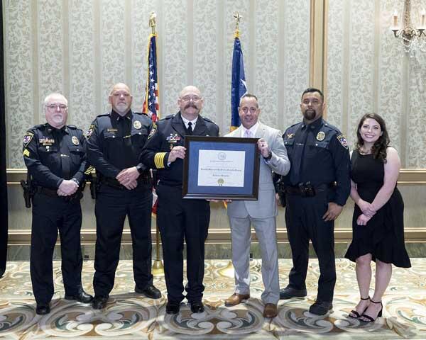 Four police officers, a gentleman, and a woman stand together smiling as they hold a large, framed award.