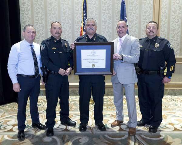 Two gentleman and three police officers stand together holding a large, framed award.