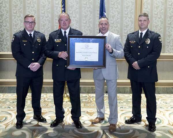 Three police officers stand smiling with a gentleman as they hold a large, framed award.