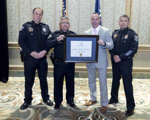 Three police officers stand next to a gentleman as they hold a large, framed award.