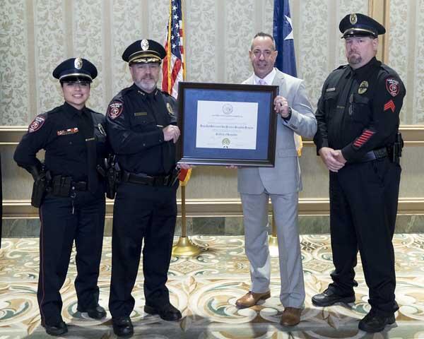 Three police officers stand next to a gentleman and smile as he holds a large award.