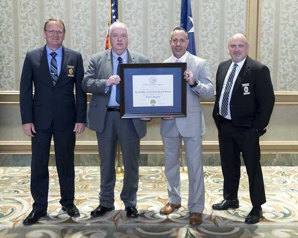 Four gentleman stand side by side, holding a large, framed award.