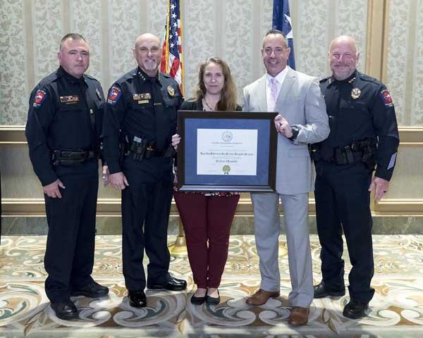 Three police officers and a gentleman stand along either side of a woman that is smiling and holding a large, framed award. 